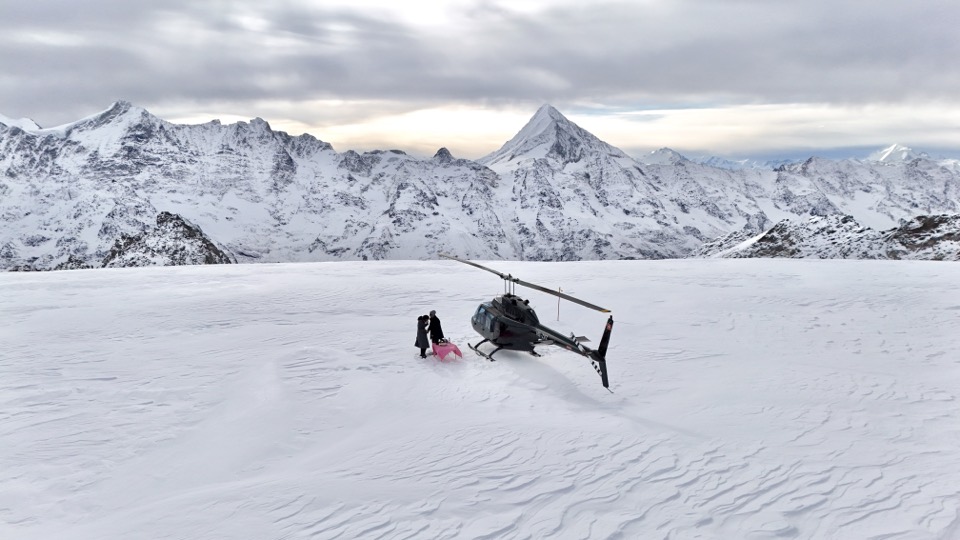 Helikopter der Airport Helicopter AHB AG auf einem Gletscher, daneben zwei Gäste an einem Apérotisch vor Bergpanorama
