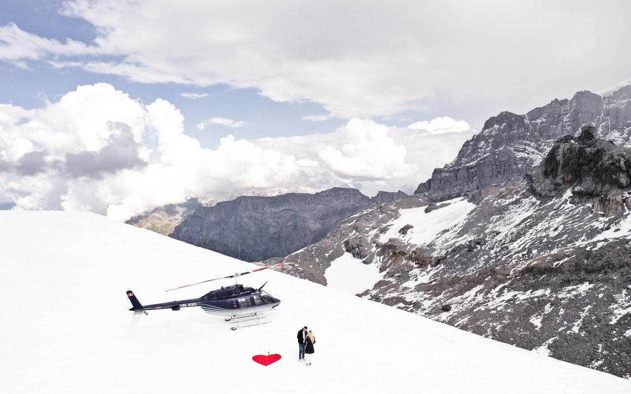 Helikopter auf einem Schneefeld, daneben ein Paar und ein rotes Herz im Schnee